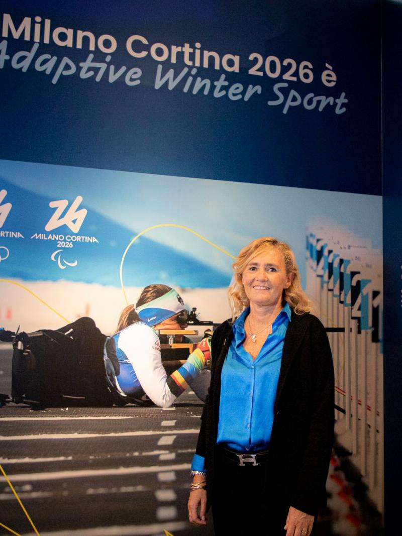A woman in business attire poses in front of a poster featuring a Para biathlon athlete in action.